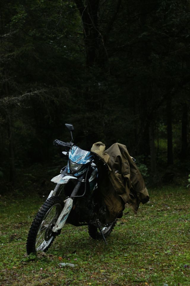 A trail motorcycle with a rider's jacket draped over it, parked in a dark forest clearing