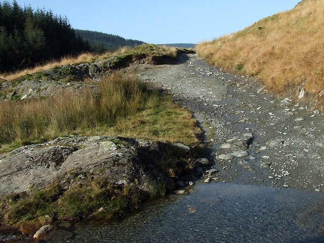 The Strata Florida byway in Powys, a rocky track with loose stone and a shallow stream crossing between grass banks and conifer forest