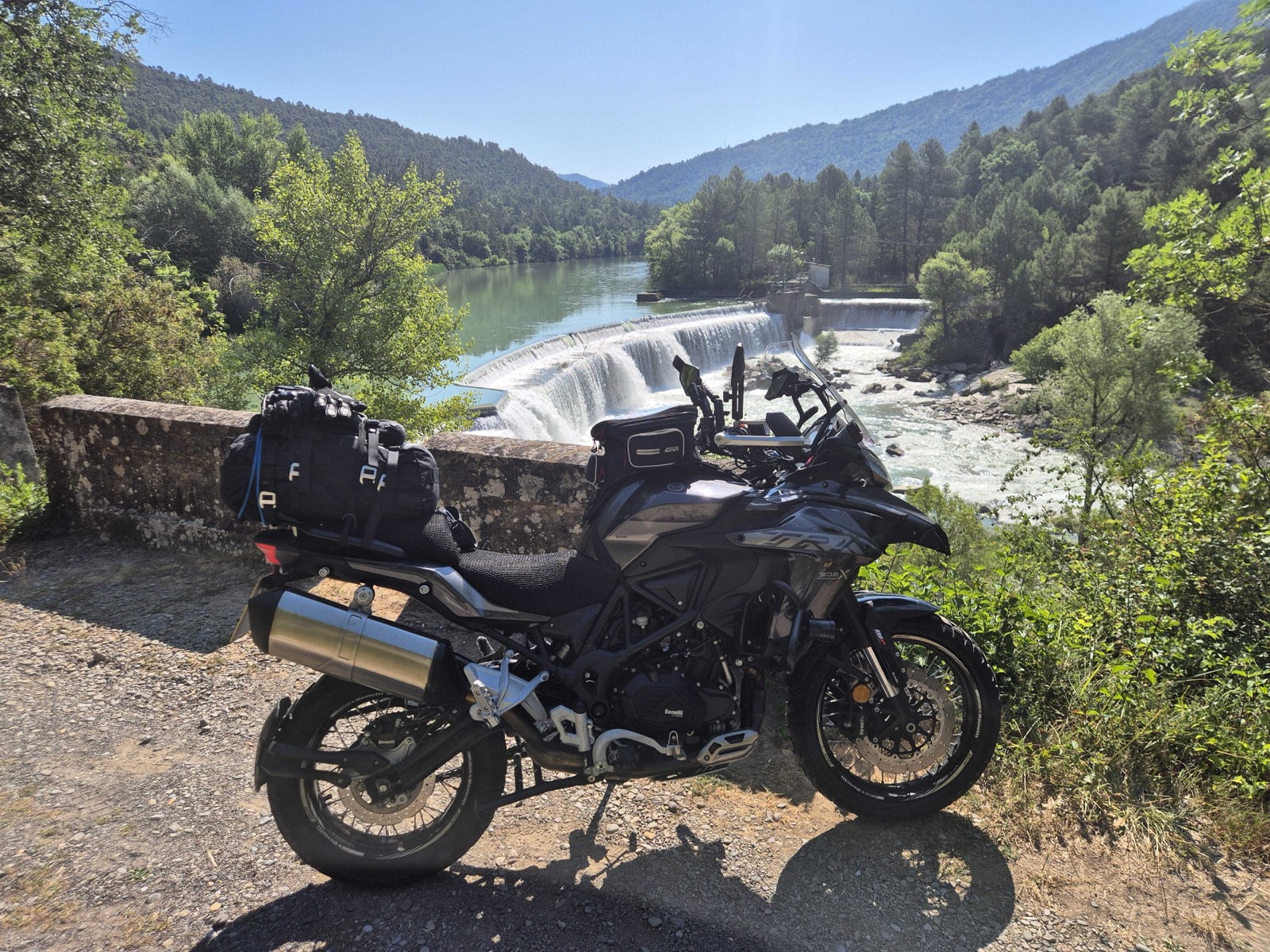 Benelli TRK 502 X parked above a weir on a river in the French Pyrenees, fully loaded with touring luggage