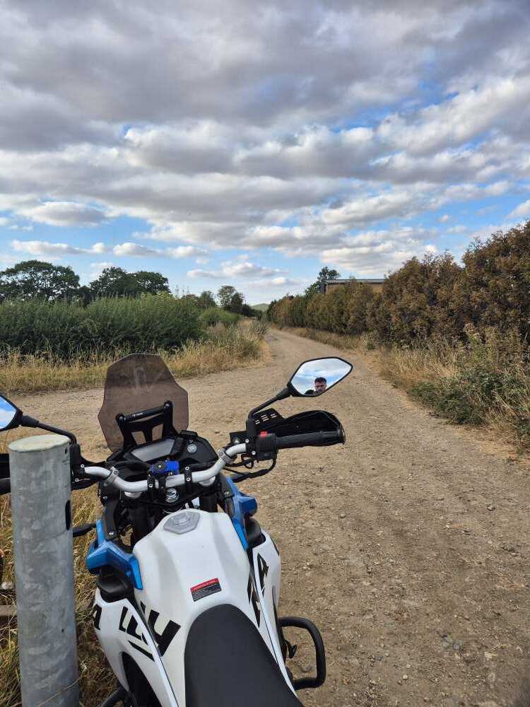 Looking down a gravel byway from the handlebars of a motorcycle, with a hedge on one side and fields on the other