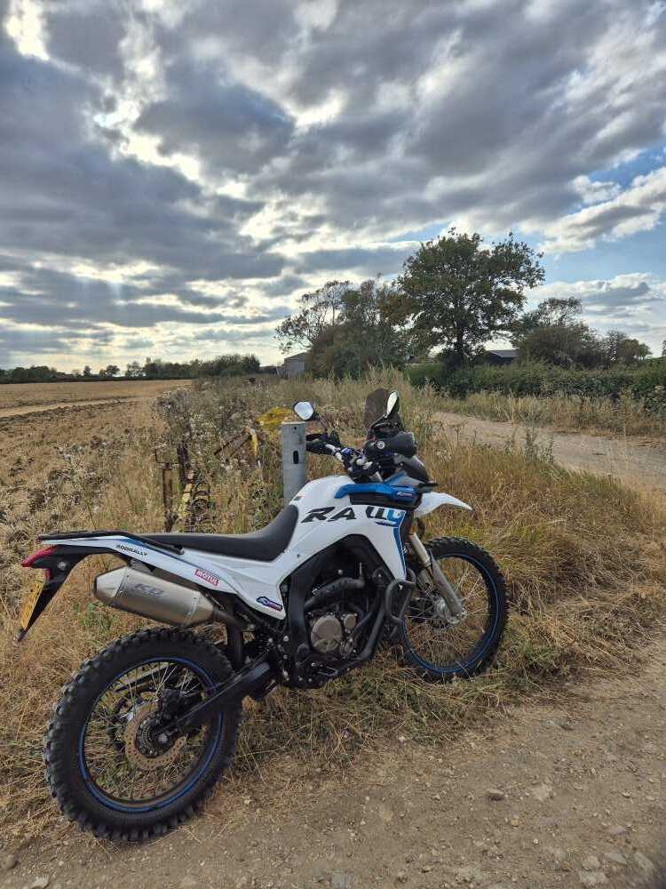 A white Voge 300 Rally parked on the grass verge of Cross End Lane under a big cloudy sky