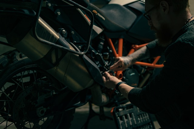 a rider working on the exhaust and engine of an orange-framed KTM with hand tools